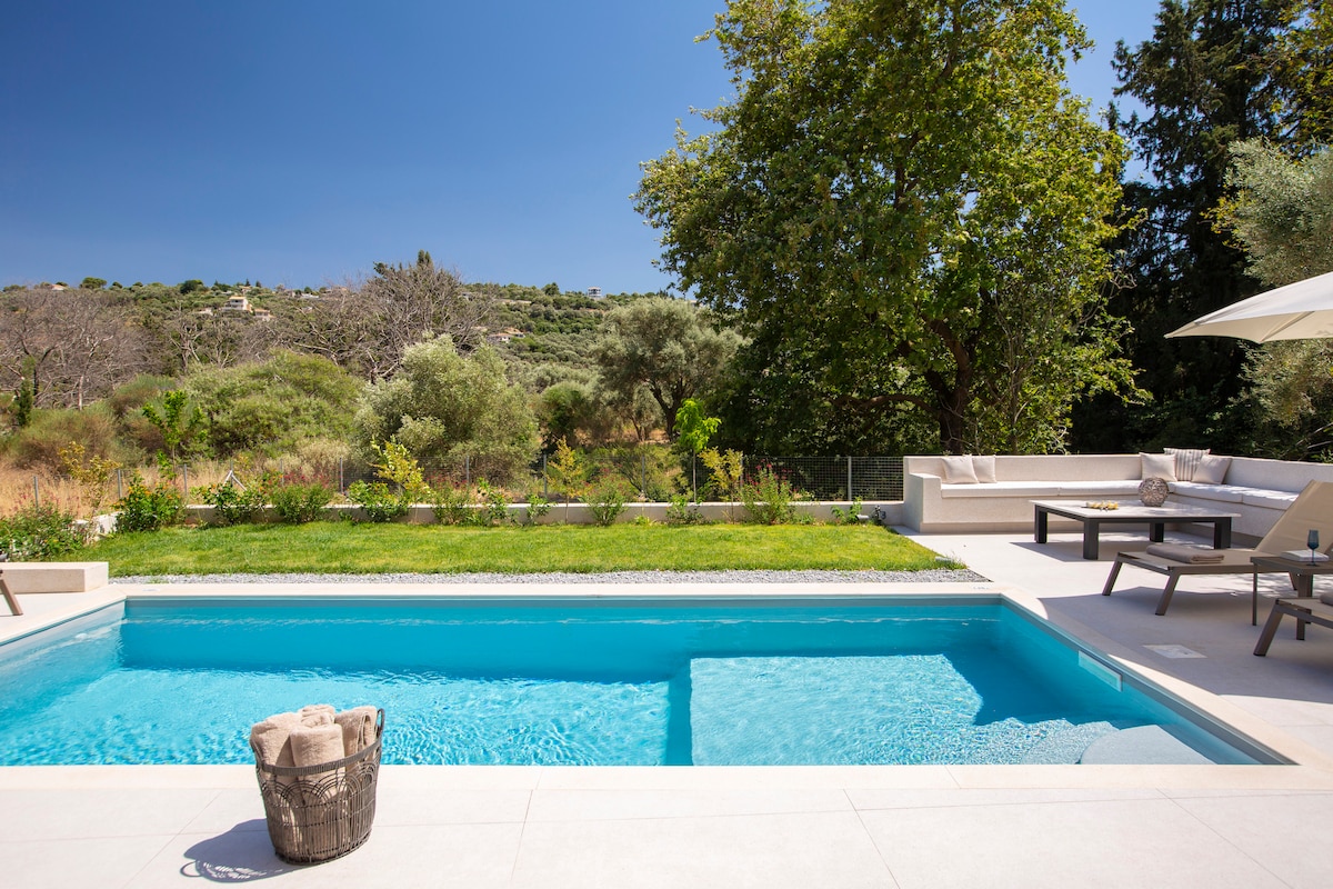 A private pool glistens under the sun, surrounded by a spacious terrace. Sun loungers and an outdoor seating area provide places to relax and enjoy nature. Lush green trees and plants frame the background, enhancing the peaceful setting of the villa.
