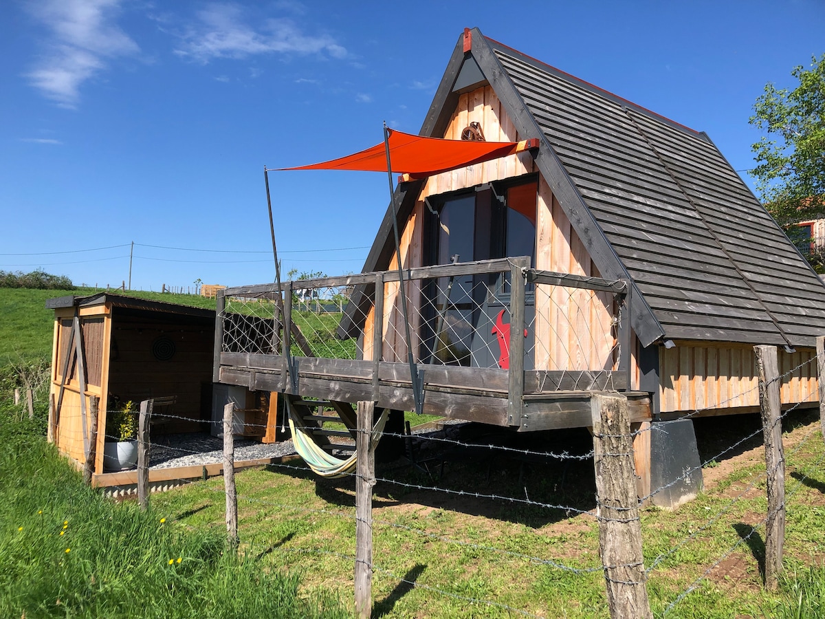 A charming A-frame cabin is featured in an outdoor setting, showcasing a spacious terrace with a hammock. Bright blue skies enhance the natural surroundings, and a red sunshade provides a pop of color. The cabin's wooden exterior complements the pastoral landscape.