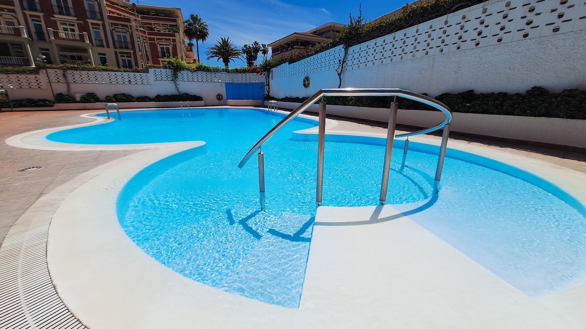 A inviting pool area is visible, featuring a unique shape and surrounded by a light-colored deck. The clear blue water reflects sunlight, and a gradual staircase leads into the pool, offering easy access. Lush greenery along the walls enhances the serene atmosphere.