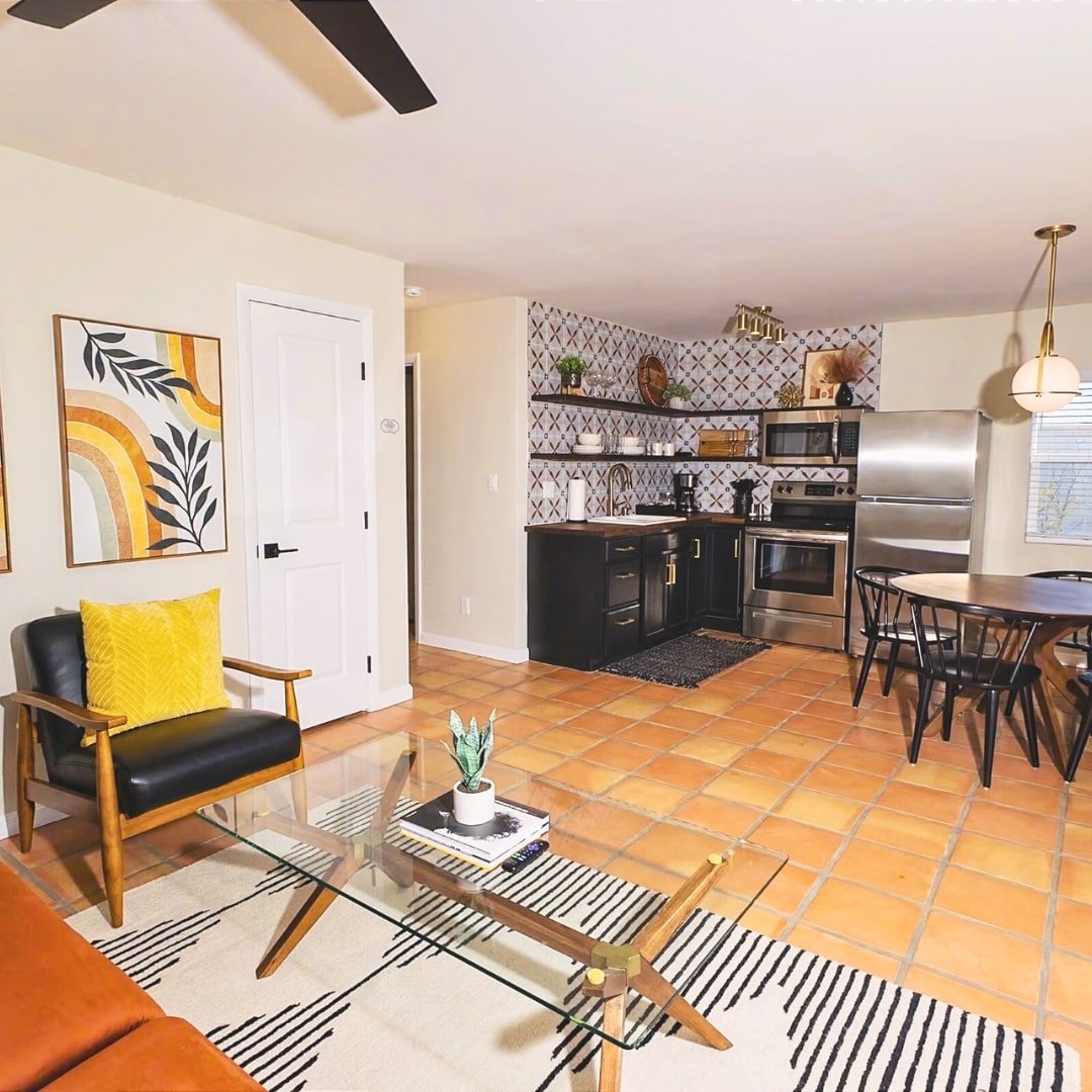 A stylish kitchen area is featured, showcasing modern appliances and geometric tile accents. A comfortable seating area includes a black armchair with a plush yellow cushion. A dining table with black chairs is visible, complementing the warm tile floor.