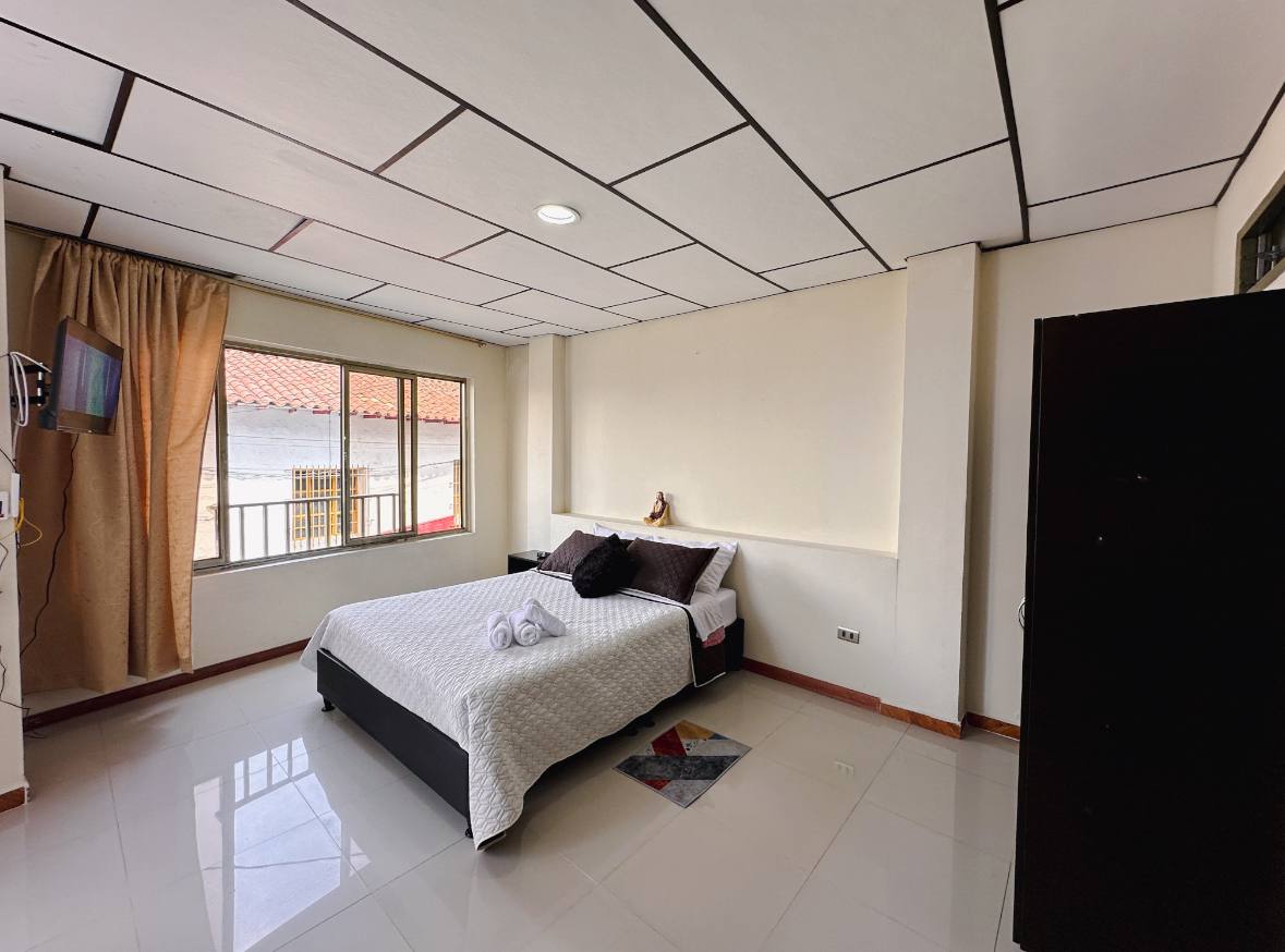 A spacious bedroom is presented with a large bed covered in a light quilt. Natural light filters through two large windows, creating a bright environment. A television is mounted on the wall, and a dark wardrobe stands against one side, complementing the room's clean design.