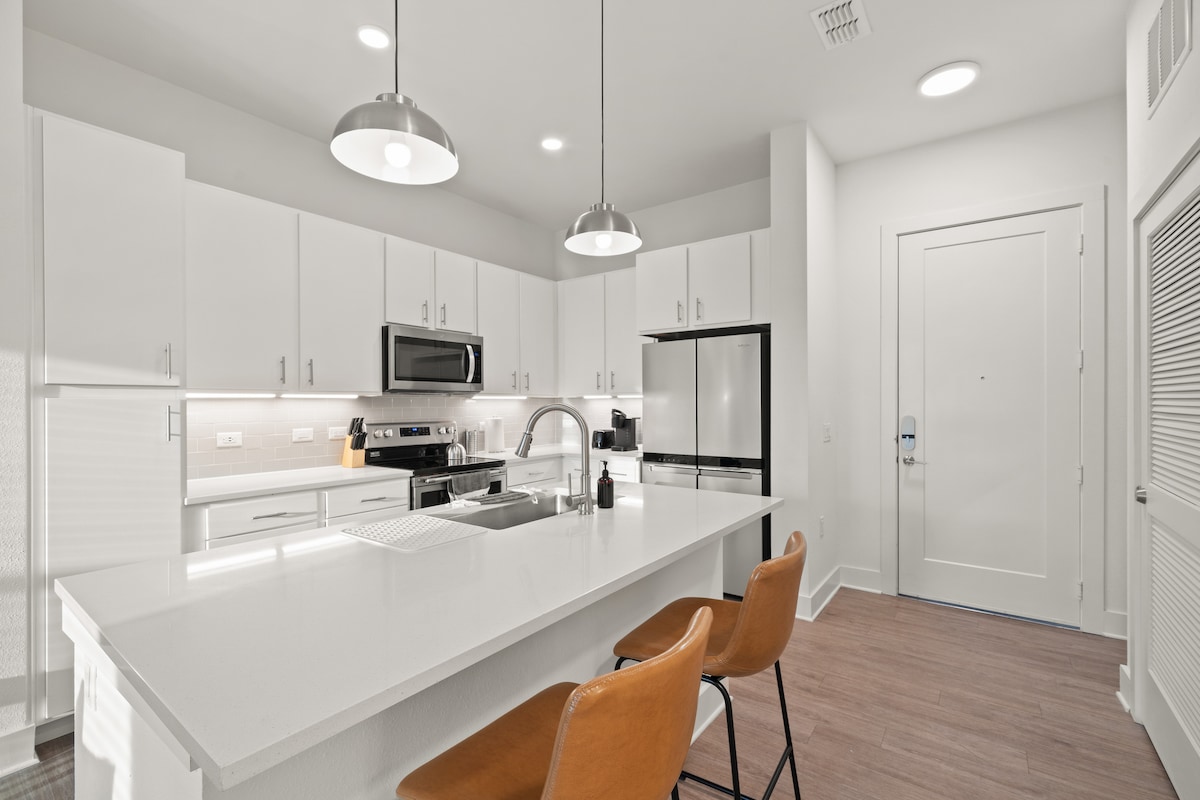 A modern kitchen showcases sleek white cabinetry and stainless steel appliances. A spacious island is complemented by two brown bar stools. Soft LED lighting illuminates the area, and the entrance door is visible in the background, enhancing the open layout.