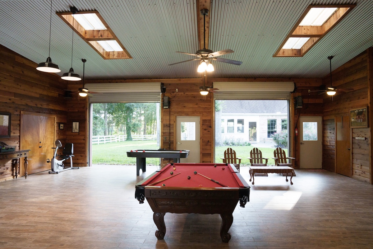 A rustic party barn interior features wooden walls and ceiling. A pool table and air hockey table are prominently displayed. Natural light filters through skylights and large windows, creating a bright atmosphere while showcasing views of the surrounding woods.