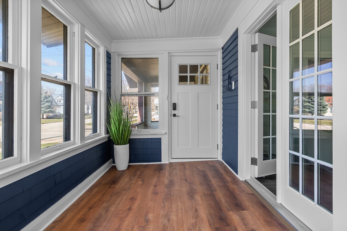 An enclosed porch is highlighted with large windows allowing natural light. A decorative potted plant adds a touch of greenery. The space features a wood floor and a door leading outside, enhancing accessibility to the outdoor area.