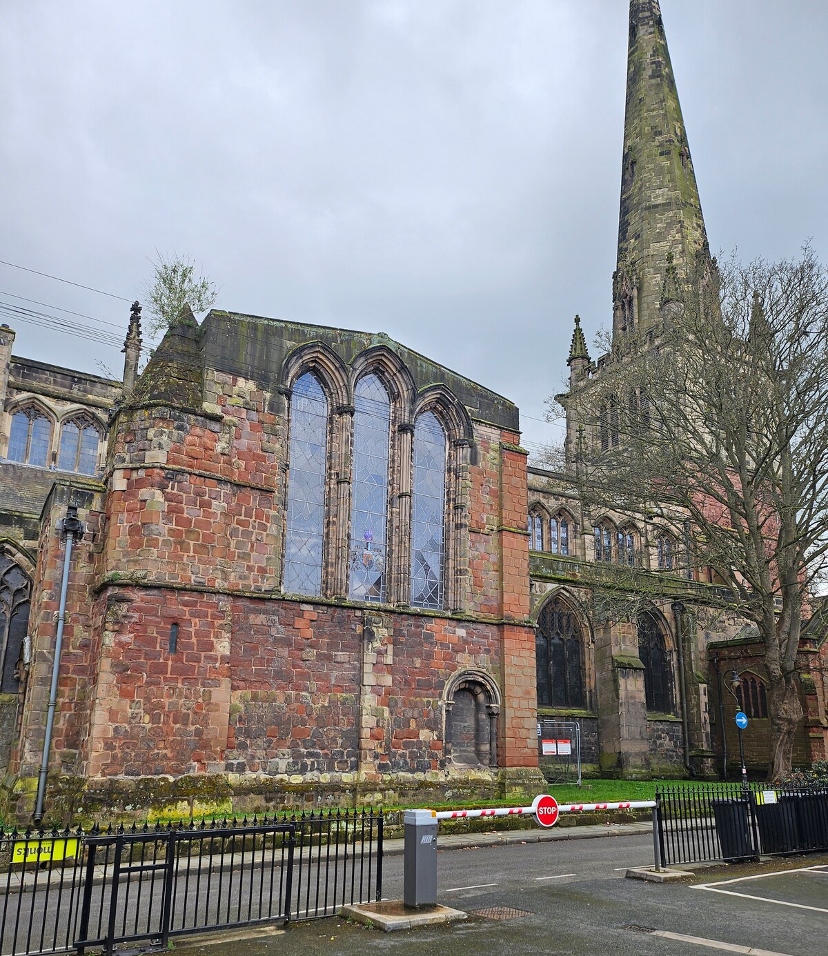 An historic building features intricate stonework with large stained glass windows. The tall spire rises above the structure, surrounded by trees and a low black fence. Nearby road signs and barriers indicate traffic regulations in the area, contributing to the scene's urban context.