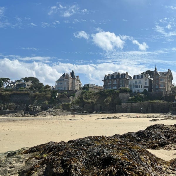 The image captures a beach scene with soft sand and scattered rocks covered in seaweed. In the background, charming historical buildings with distinctive architecture rise from the cliff, under a clear blue sky with scattered clouds.