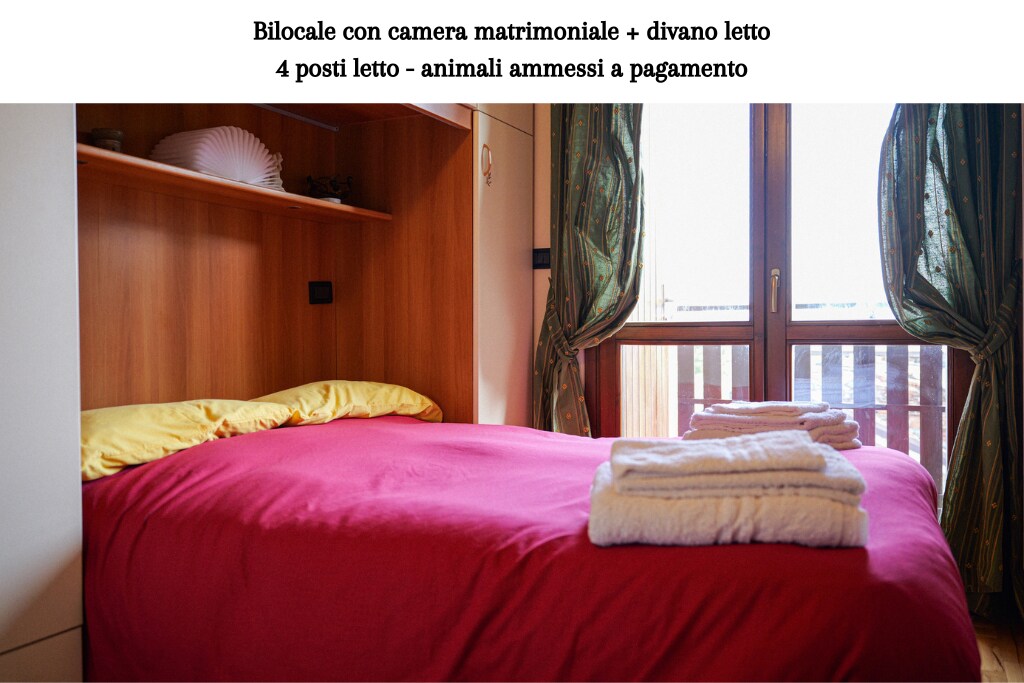 A cozy sleeping area features a spacious bed covered with a vibrant red blanket. Soft white towels are neatly arranged at the foot of the bed. Natural light enters through a window dressed in patterned curtains, illuminating the warm wooden accents in the room.