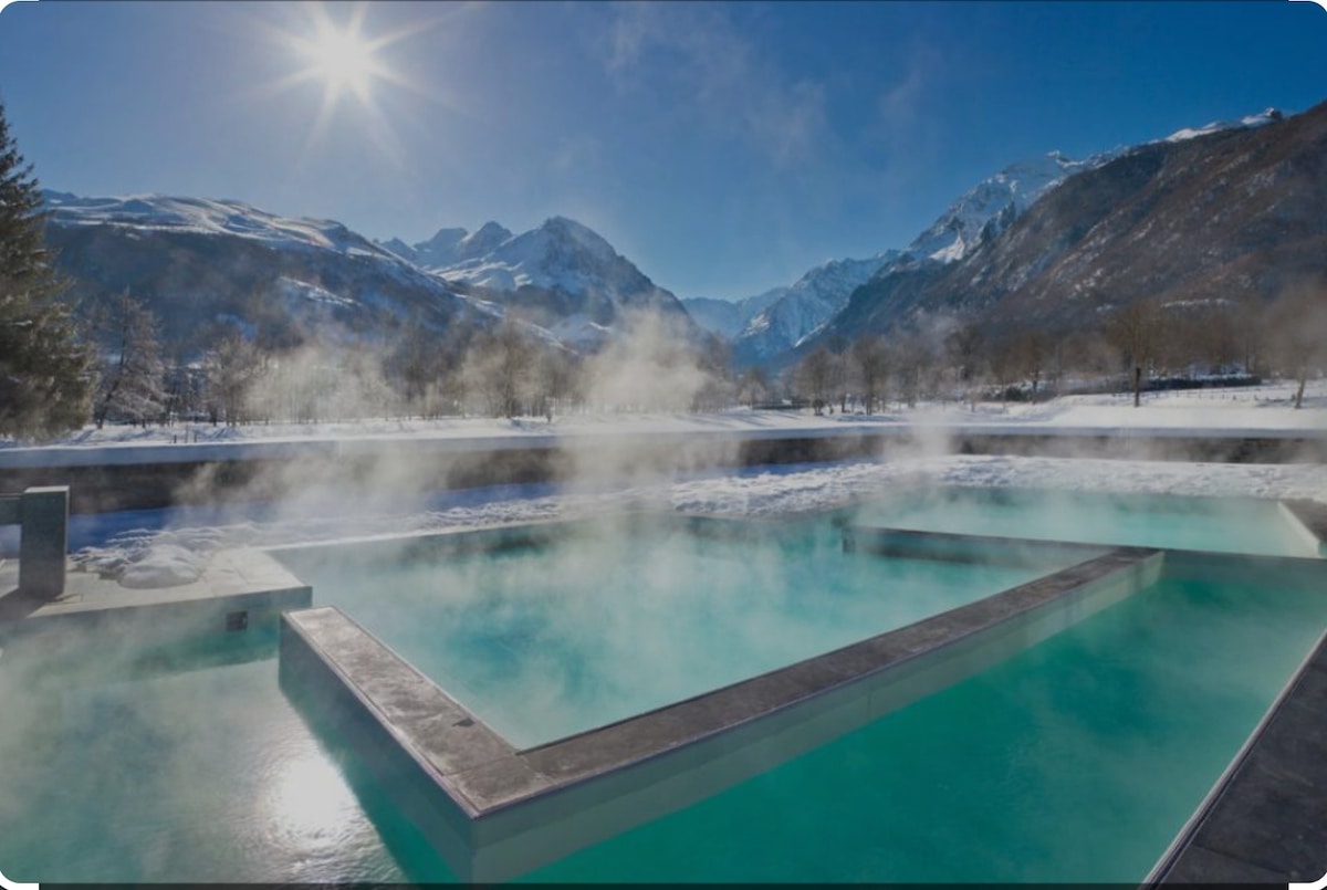 An outdoor thermal pool is surrounded by snow-covered mountains, with steam rising gently from the water. Bright sunlight reflects off the pool's surface, creating a serene and inviting scene in a winter landscape.