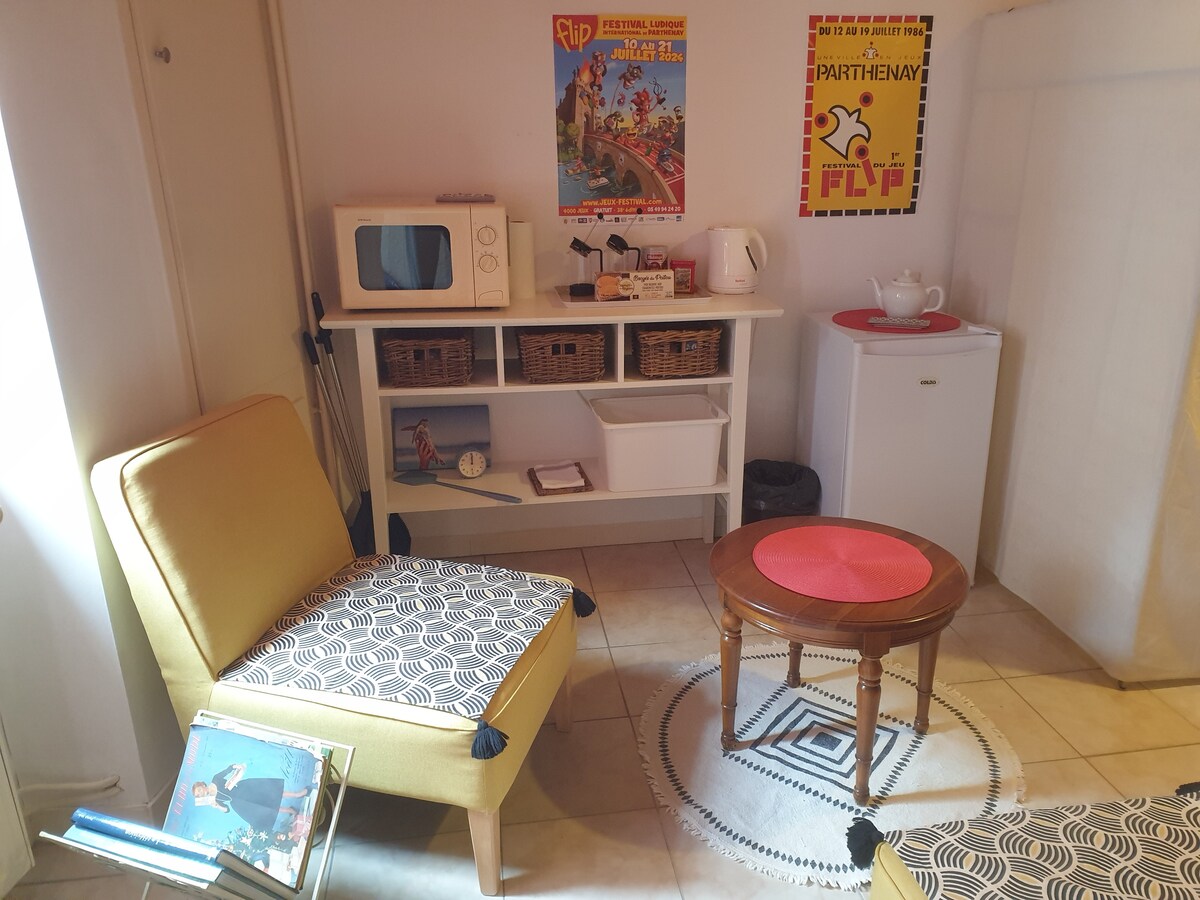 A cozy corner of the studio features a bright yellow chair with a patterned cushion, complemented by a round wooden table. A microwave and a mini-fridge are situated on a shelf, along with decorative baskets. Vintage posters adorn the walls, adding character to the space.