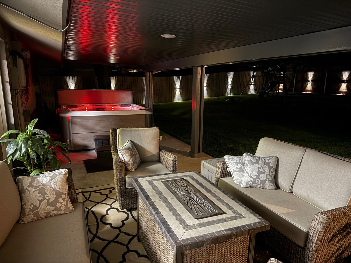 An outdoor seating area is featured under a covered porch, furnished with a light-colored couch and a patterned table. In the background, a hot tub is illuminated with soft red lighting, and softly lit trees are visible beyond the yard, enhancing the serene atmosphere.