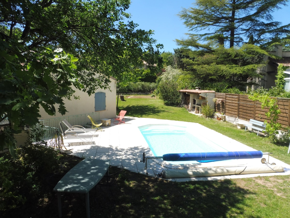 A tranquil outdoor area features a swimming pool surrounded by a lawn. Sun loungers and chairs provide seating options, while a shaded dining table offers a comfortable space for relaxation. Lush trees and a small garden enhance the privacy and serene atmosphere.
