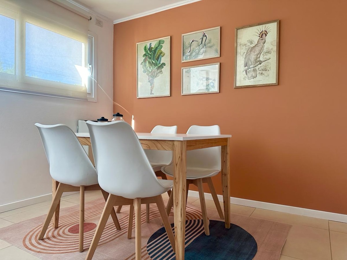 A bright dining area features a wooden table surrounded by four white chairs. Natural light filters through a window with sheer blinds, casting soft shadows. The walls are painted in a warm orange hue, complemented by framed botanical and wildlife art pieces.