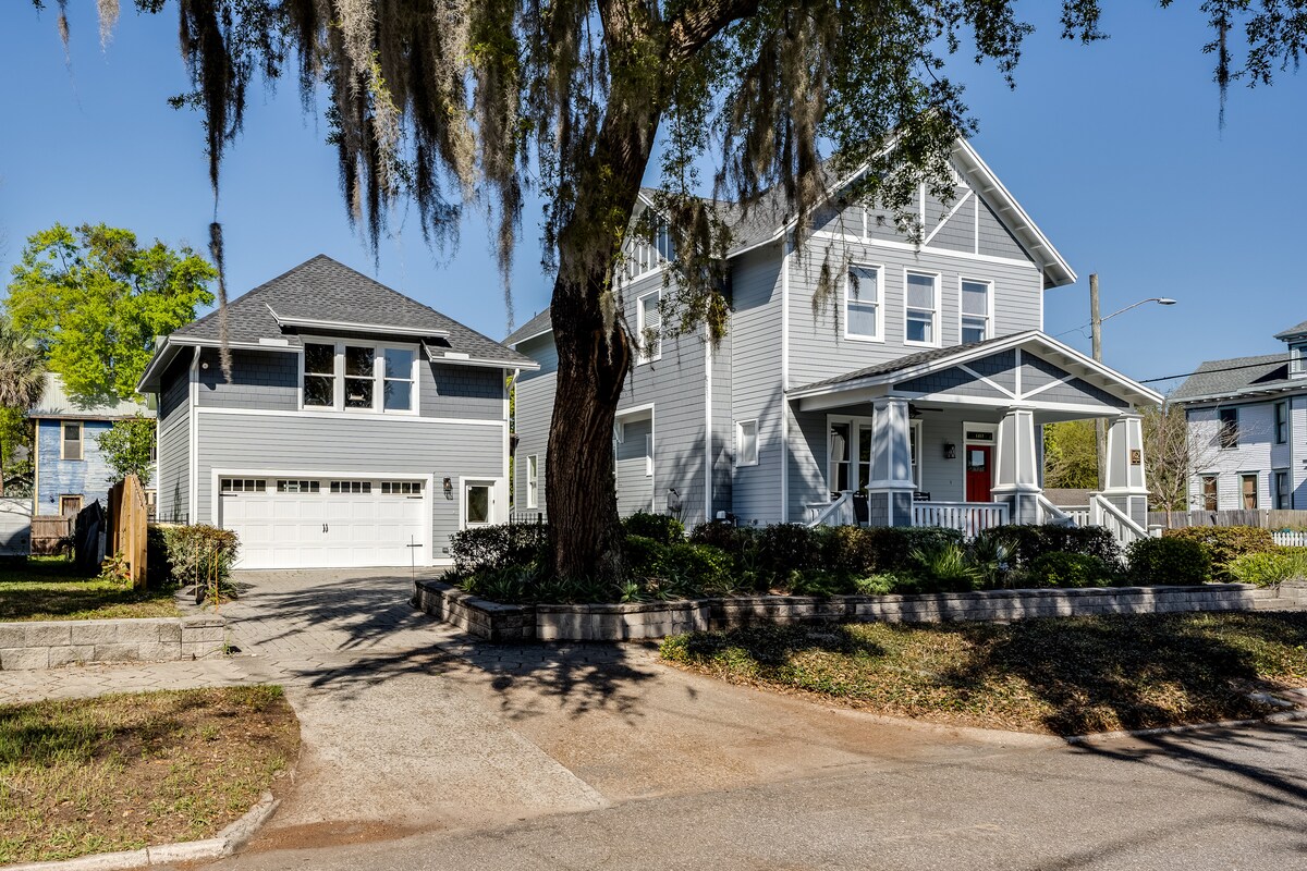 The exterior of the guesthouse is featured, showcasing a two-story, light blue structure with a steep roof. Surrounding greenery includes mature trees and manicured shrubs. A driveway leads to the garage below the guesthouse, with a peaceful street scene visible in the foreground.