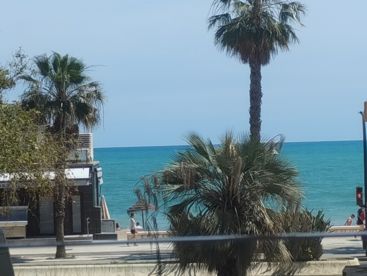 The image captures a view of the beach featuring gentle waves and turquoise water. Lush palm trees frame the scene, while a sandy shore is visible in the distance. A parasol and beach-goers can be seen, providing a sense of a lively coastal atmosphere.