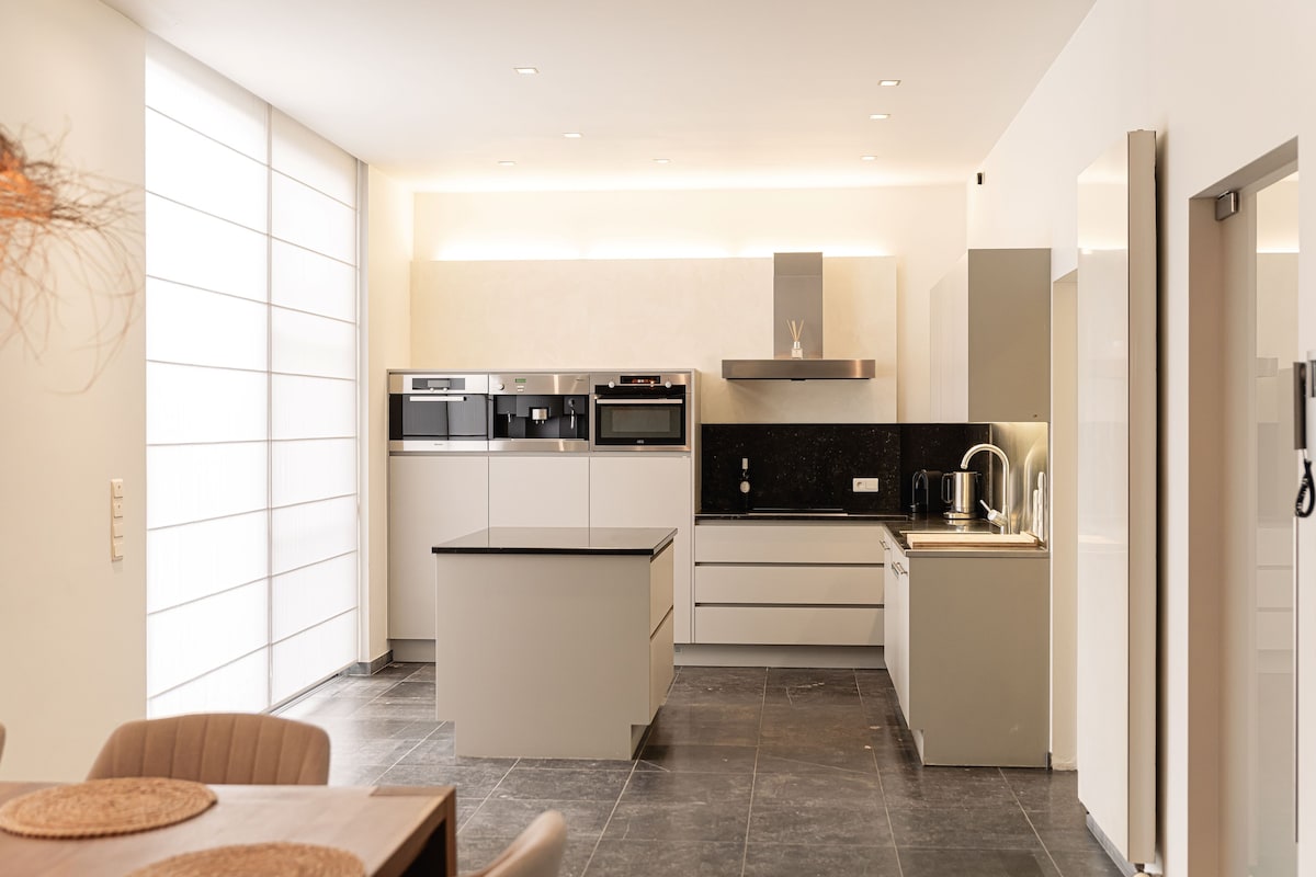 A modern kitchen features sleek cabinetry and a central island, providing ample workspace. High-end appliances are integrated seamlessly into the design. Natural light floods the space through large windows, enhancing the clean and contemporary aesthetic of the area.