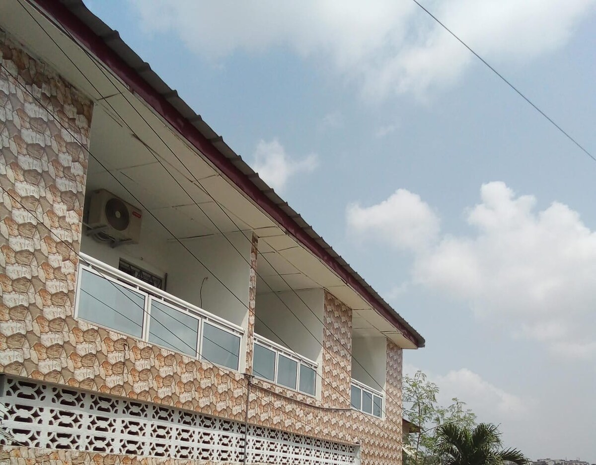 The exterior of the building showcases a two-story structure with textured wall designs. Large windows provide ample light and views, while an air conditioning unit is visible for comfort. A clear blue sky with scattered clouds serves as a backdrop.