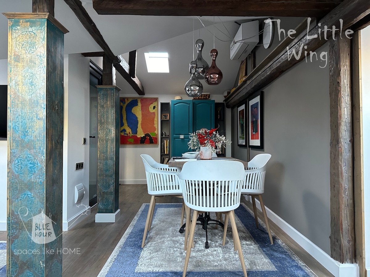 An elegantly designed dining area is featured, showcasing a round table surrounded by modern white chairs. Subtle blue hues can be seen on the walls, with artwork adding color to the space. Exposed wooden beams enhance the room's character, and natural light filters through the skylight.