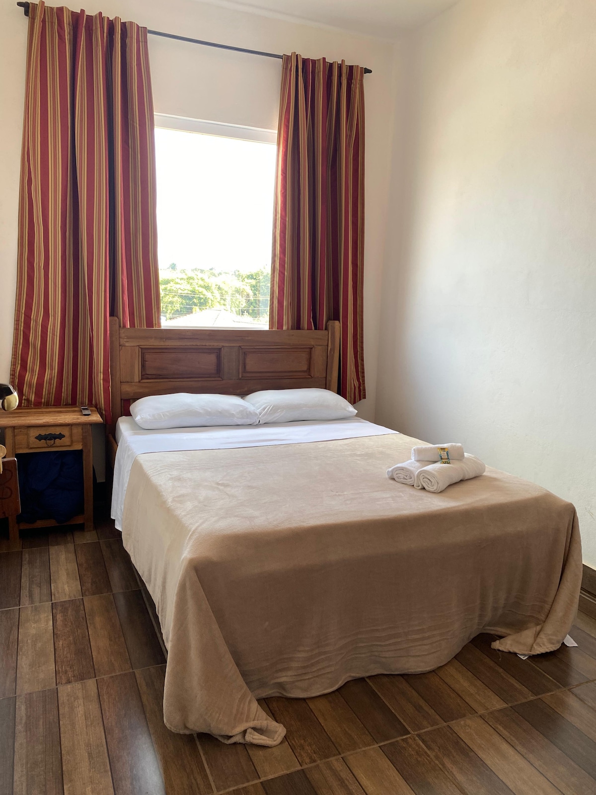A cozy sleeping area features a spacious bed with neutral bedding and neatly rolled towels. Natural light floods the room through a large window, framed by striped curtains. Wooden furnishings, including a bedside table, complement the warm atmosphere.