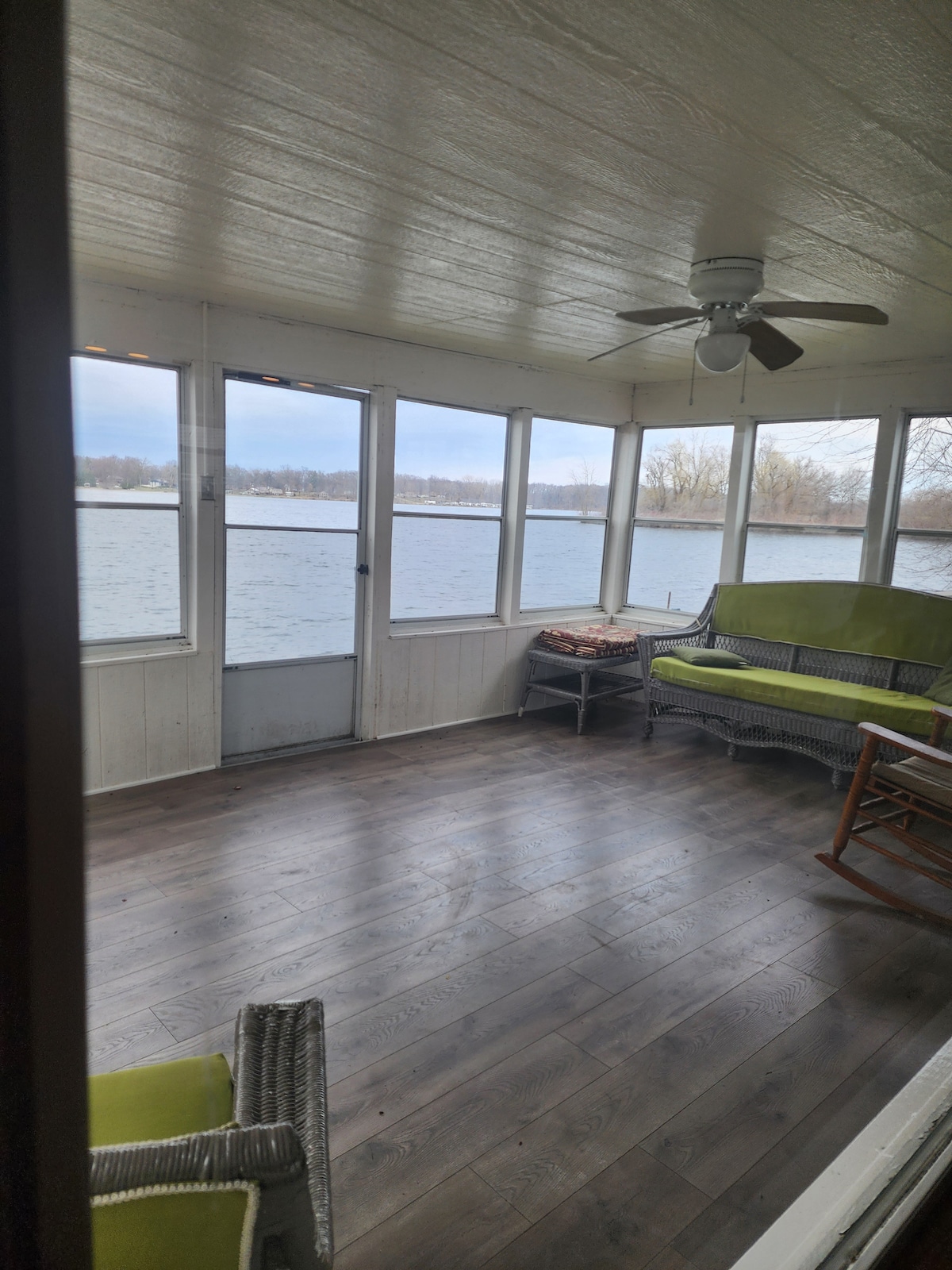 A sunroom with large windows overlooking the lake creates a bright and airy atmosphere. A green couch and a rocking chair are positioned opposite a small table, inviting relaxation. The floor is finished in dark wood, and a ceiling fan can be seen above.