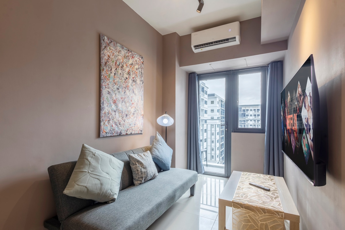 A modern living area features a spacious sofa with decorative pillows, complemented by a stylish coffee table. Natural light enters through a large window, and a wall-mounted Smart TV emphasizes entertainment options. An abstract artwork adds a pop of color to the neutral walls.