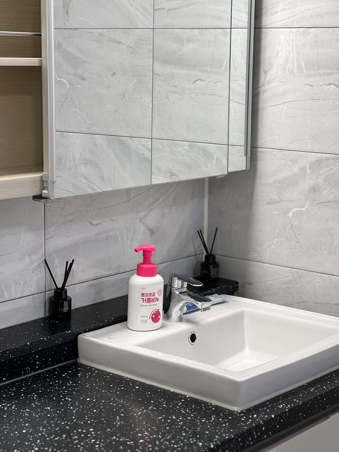 A clean bathroom countertop is visible, featuring a white sink with a modern faucet. A pink liquid soap dispenser sits atop the black speckled surface. A mirror above the sink reflects the light gray tiled walls, while decorative reed diffusers are positioned on either side of the faucet.