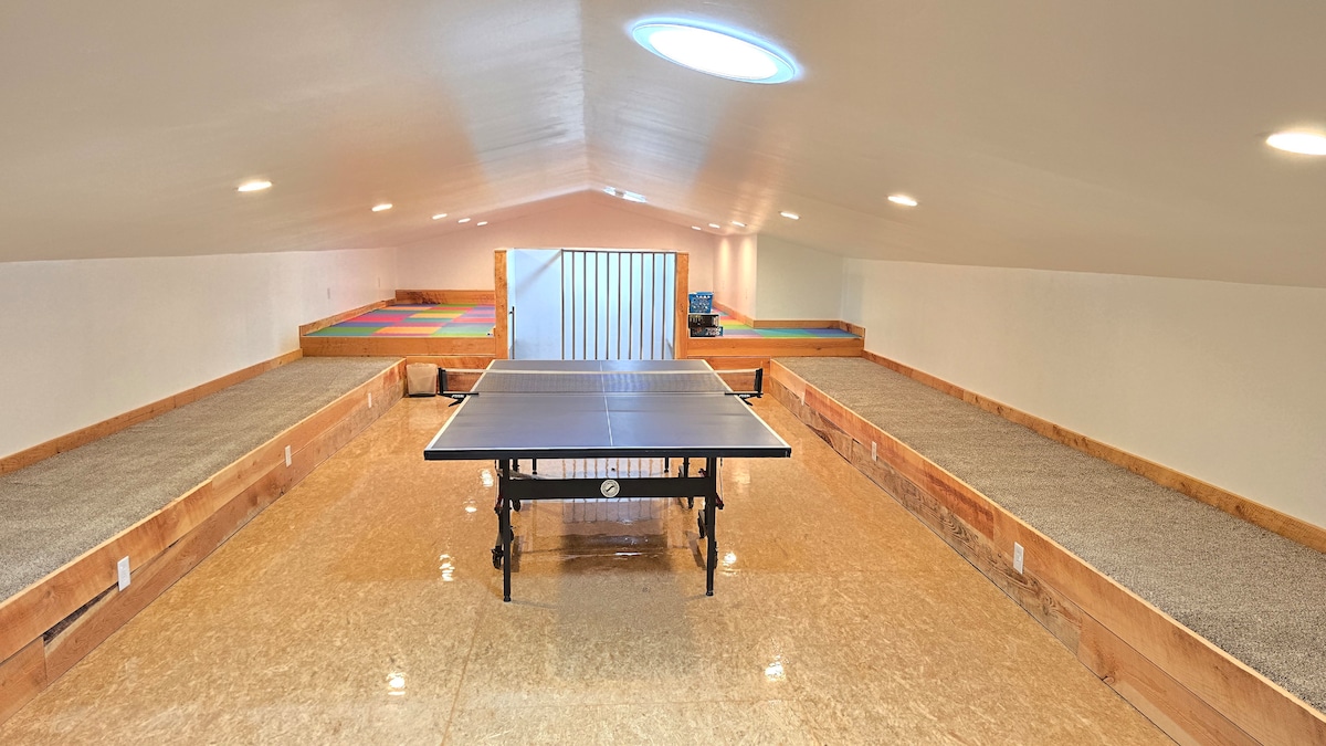 A spacious game room is featured on the upper level, showcasing a ping pong table at the center. Surrounding areas are fitted with cushioned, colorful mats, providing additional space for activities. Soft lighting from recessed fixtures enhances the bright, open atmosphere.