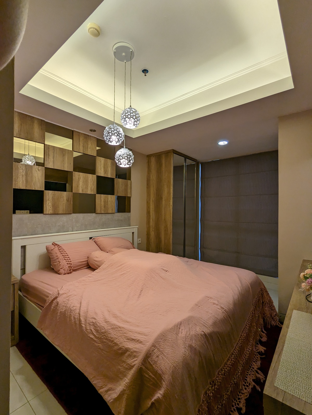 The bedroom features a spacious bed draped in pink bedding, complemented by two pillows and bolsters. Soft lighting is provided by pendant fixtures above. Natural light filters through sheer window coverings, enhancing the modern decor of the wood panel accent wall.