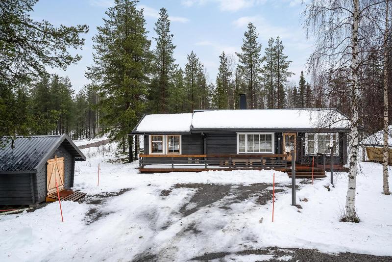 A cozy cabin is nestled among tall trees, surrounded by a blanket of snow. The building features a welcoming porch that extends across the front, while a smaller outbuilding is visible nearby. A clear driveway provides access from the road.