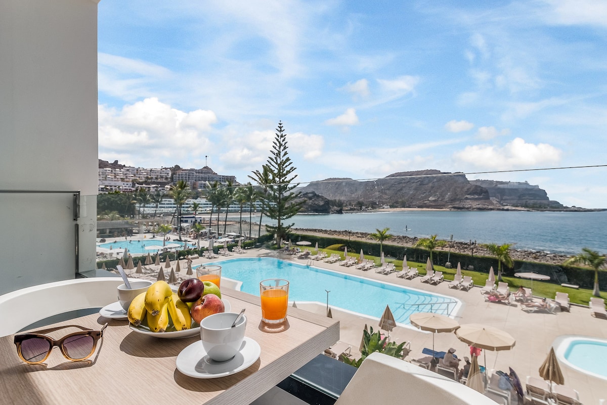A dining table on the terrace is set with a bowl of fresh fruit, a cup, and a glass of orange juice. The view features a large community pool surrounded by palm trees and scenic ocean vistas under a bright sky.