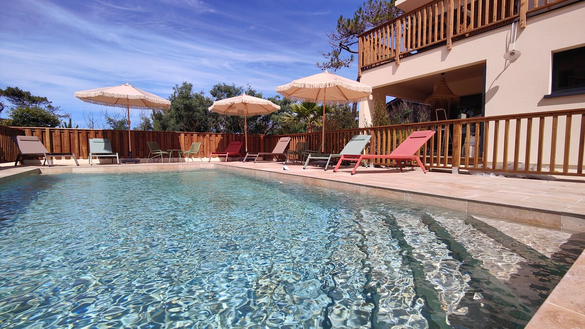 A serene pool area is featured, surrounded by comfortable lounge chairs and shaded by umbrellas. The water sparkles in the sunlight, and the smooth stone deck complements the natural landscape of trees in the background, creating a tranquil outdoor setting.