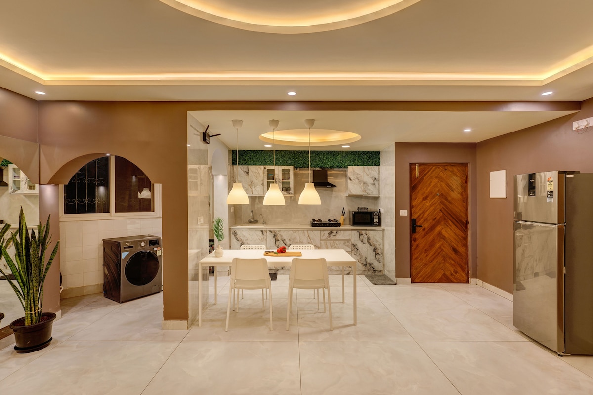 The fully equipped kitchen features sleek marble countertops and modern appliances, including a refrigerator and washing machine. A dining table with white chairs is centrally located, while warm lighting enhances the spacious feel of the area. Decorative plants add a touch of greenery to the room.