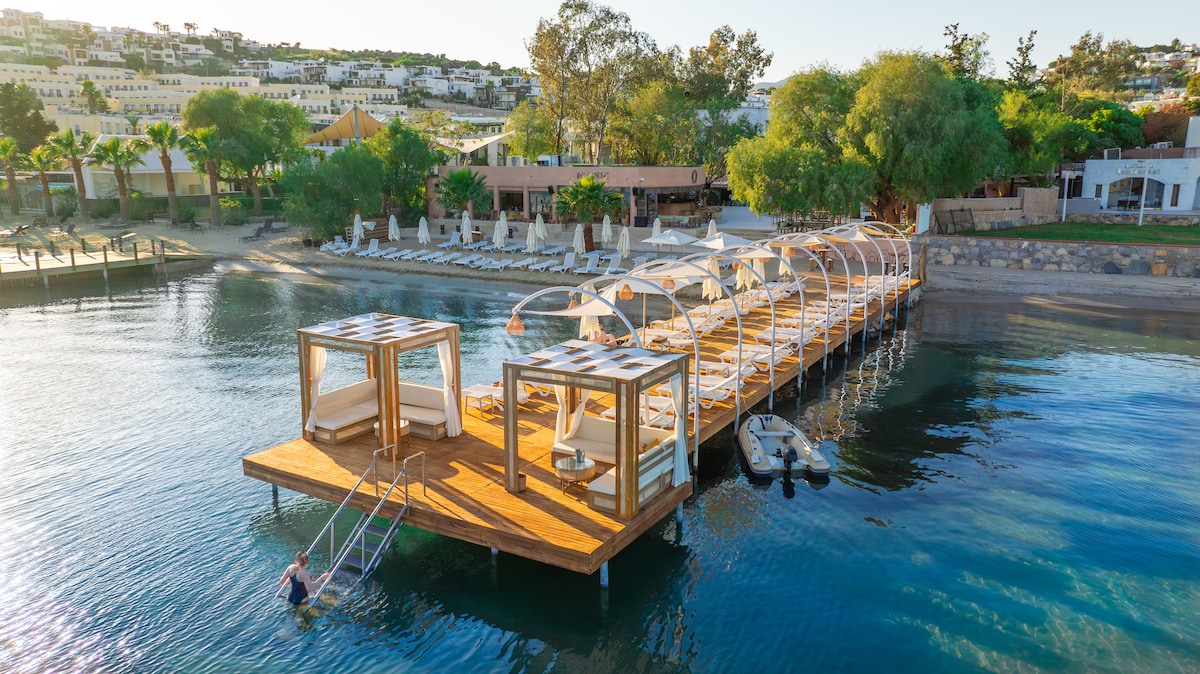 A wooden dock extends over calm waters, featuring cushioned loungers and shaded canopies. Steps lead into the water, surrounded by tropical greenery and a sandy beach area. In the background, a restaurant building and additional lounge chairs are visible along the shoreline.