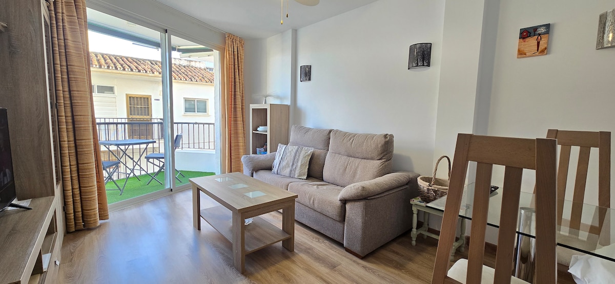 A comfortable living area features a spacious sofa and a coffee table, with a TV positioned on a wooden console. Large glass doors open to a terrace, allowing natural light to enhance the room. Dining chairs surround a glass table, creating a welcoming space.