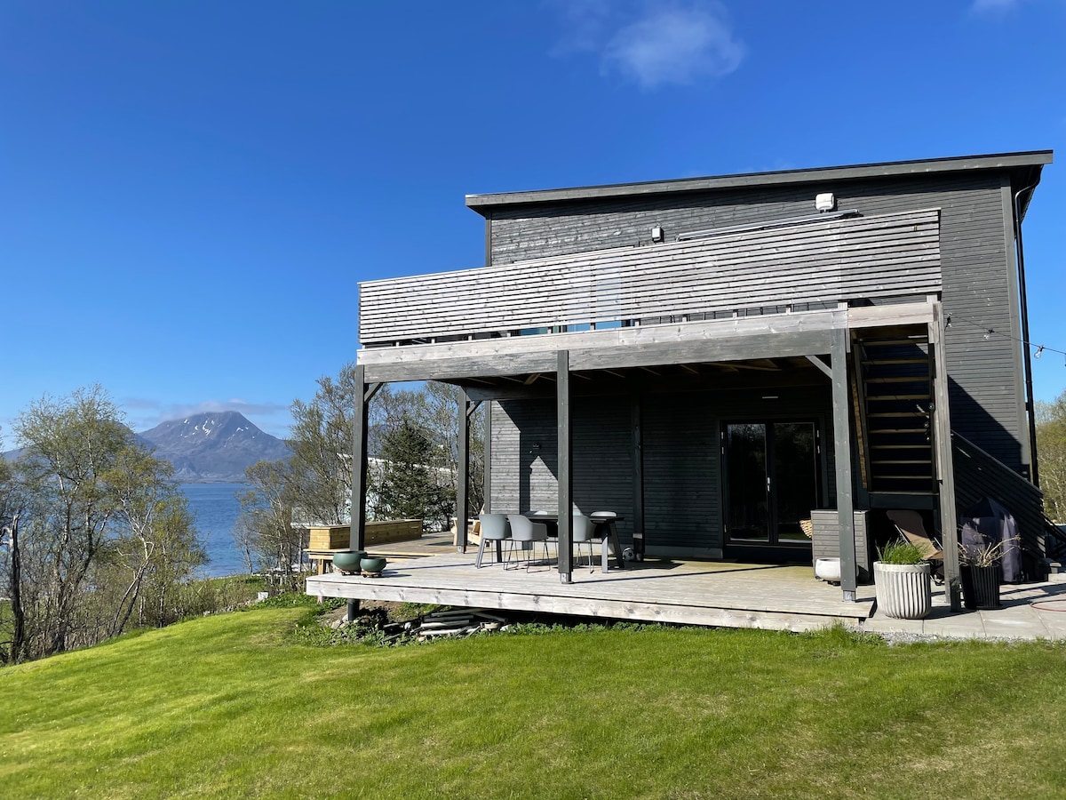A modern wooden house is depicted, featuring a large deck with outdoor seating overlooking a scenic view of the water and nearby mountains. The expansive grassy area surrounds the home, providing a sense of openness and connection to nature.