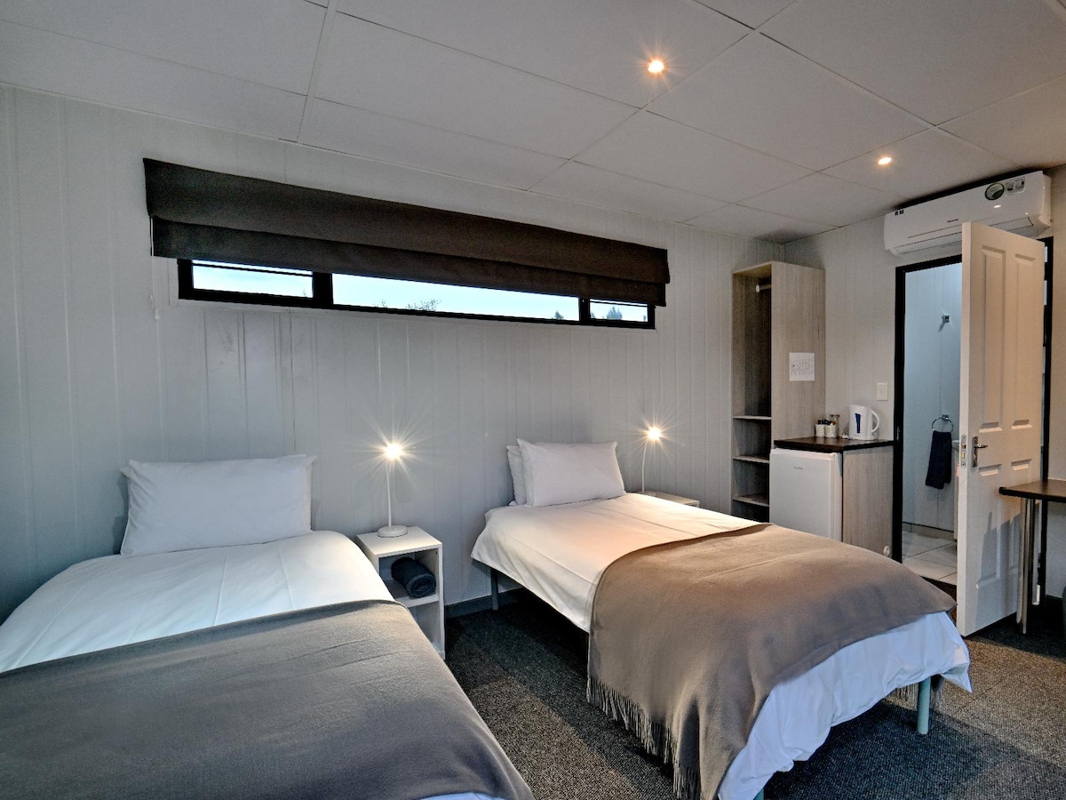 A comfortable twin room is seen, featuring two spacious beds with neutral-toned bedding. Each bed is accompanied by a modern bedside lamp. A kitchenette area is visible, complete with a bar fridge and tea/coffee station. The en-suite bathroom entrance can be seen in the background.