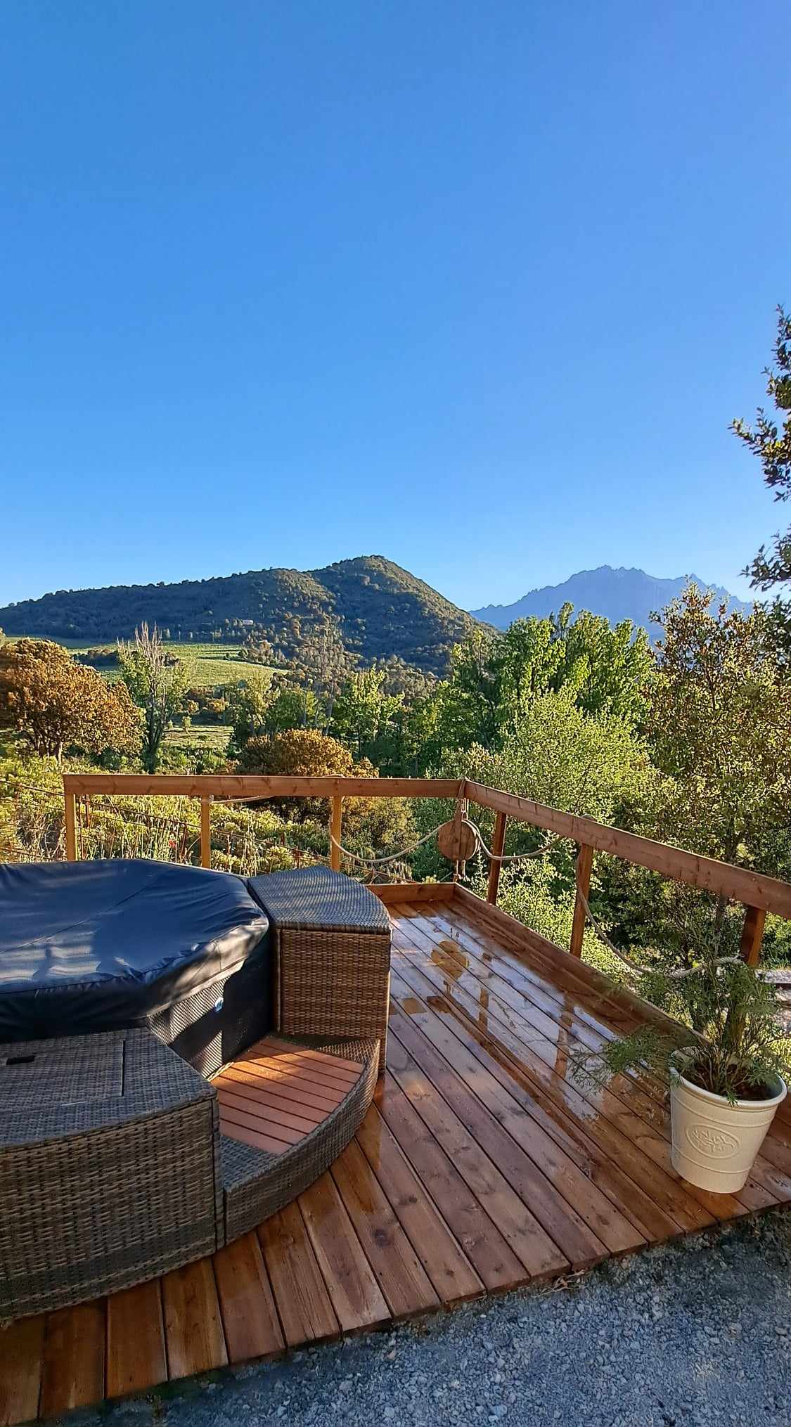 A wooden deck is showcased, featuring a hot tub surrounded by a circular seating area. Lush greenery and rolling mountains create a serene backdrop under a clear blue sky. The deck reflects sunlight, highlighting its inviting and well-maintained appearance.