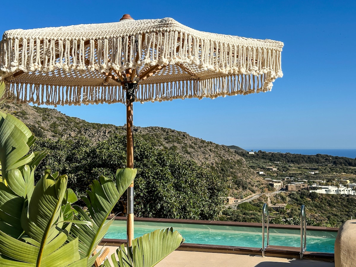 A woven umbrella offers shade by the edge of a tranquil pool, surrounded by lush greenery. The scenic hillside and distant sea are visible in the background, contributing to a serene outdoor environment.