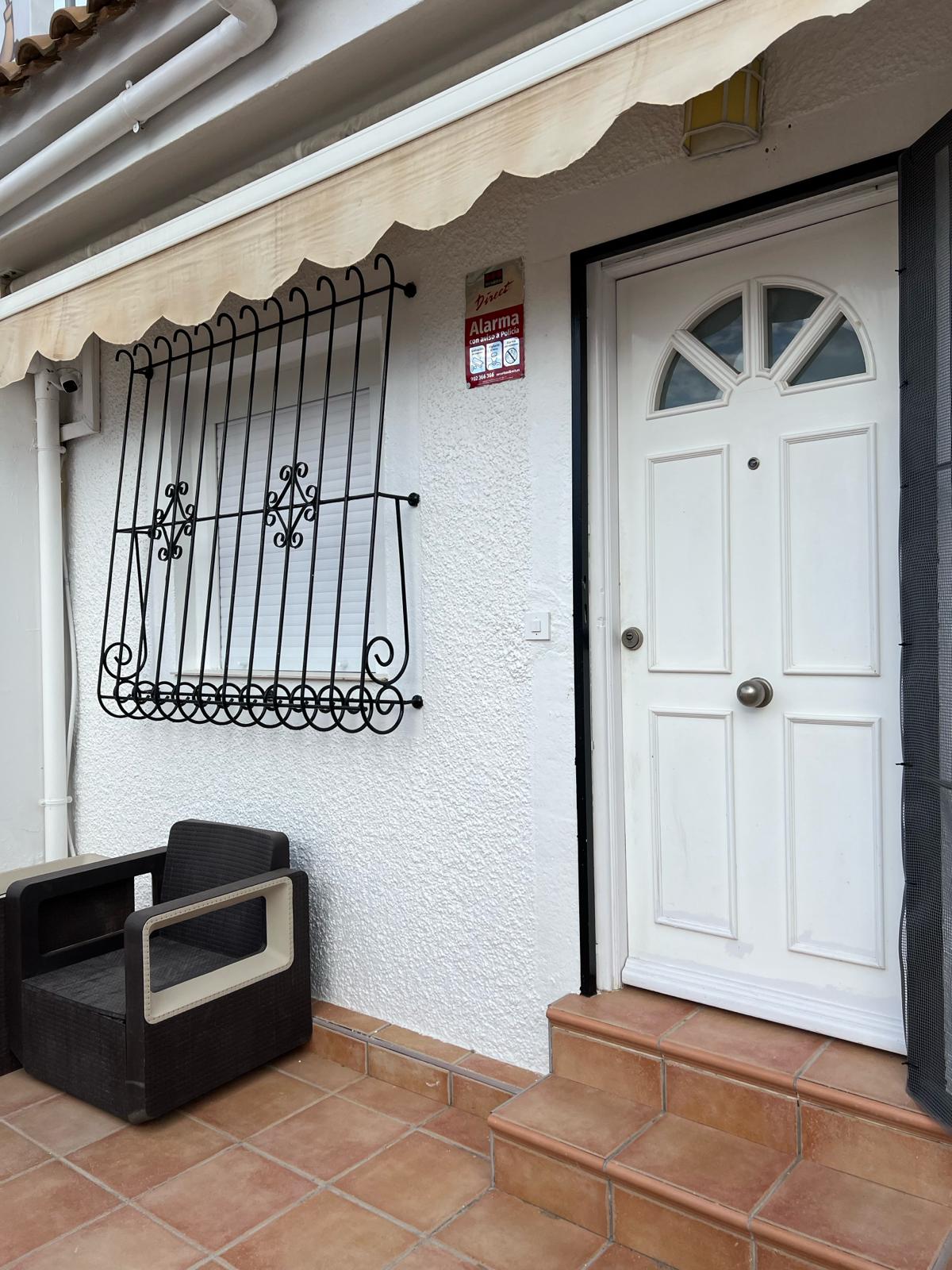 A white front door with a decorative design is flanked by a comfortable seating area featuring two dark wicker chairs. Above the door, a security grill adds an element of safety, and ceramic tiles cover the ground in a warm tone, complementing the overall entrance.