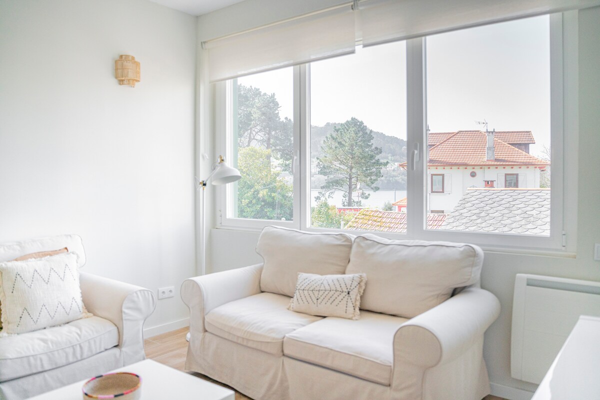 A light-filled living area features a cozy white sofa adorned with decorative cushions. Large windows provide views of the surrounding landscape, including distant hills and nearby rooftops. Natural light enhances the bright, airy atmosphere of the space.