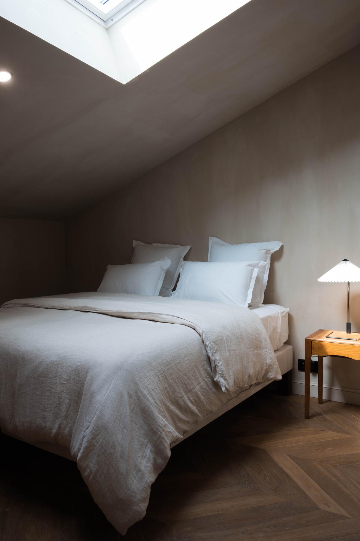 A serene bedroom features a spacious bed adorned with crisp, white linens, complemented by two soft pillows. A minimalist bedside table holds a stylish lamp, while natural light filters through a skylight, illuminating the warm wooden floor.