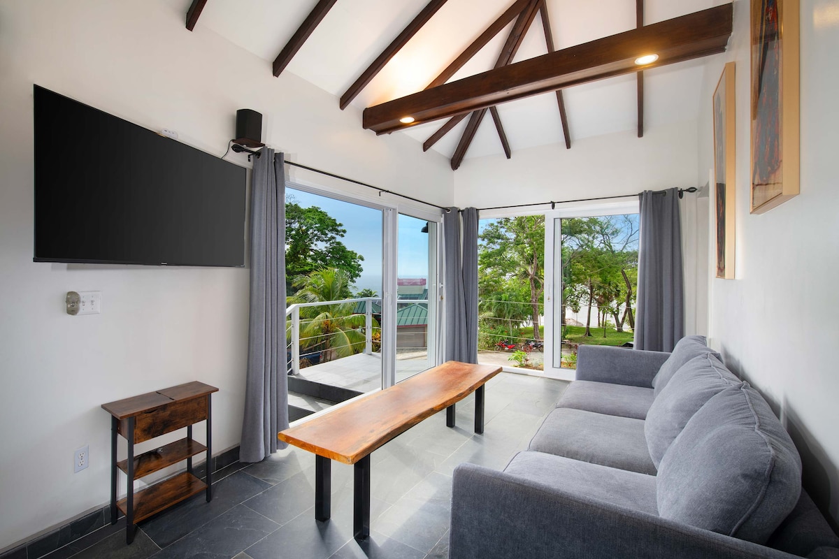 The spacious living area features a comfortable grey sofa, a modern wooden coffee table, and a large wall-mounted television. Floor-to-ceiling glass doors open to a serene outdoor view, enhancing the bright atmosphere created by natural light streaming in through the curtains.
