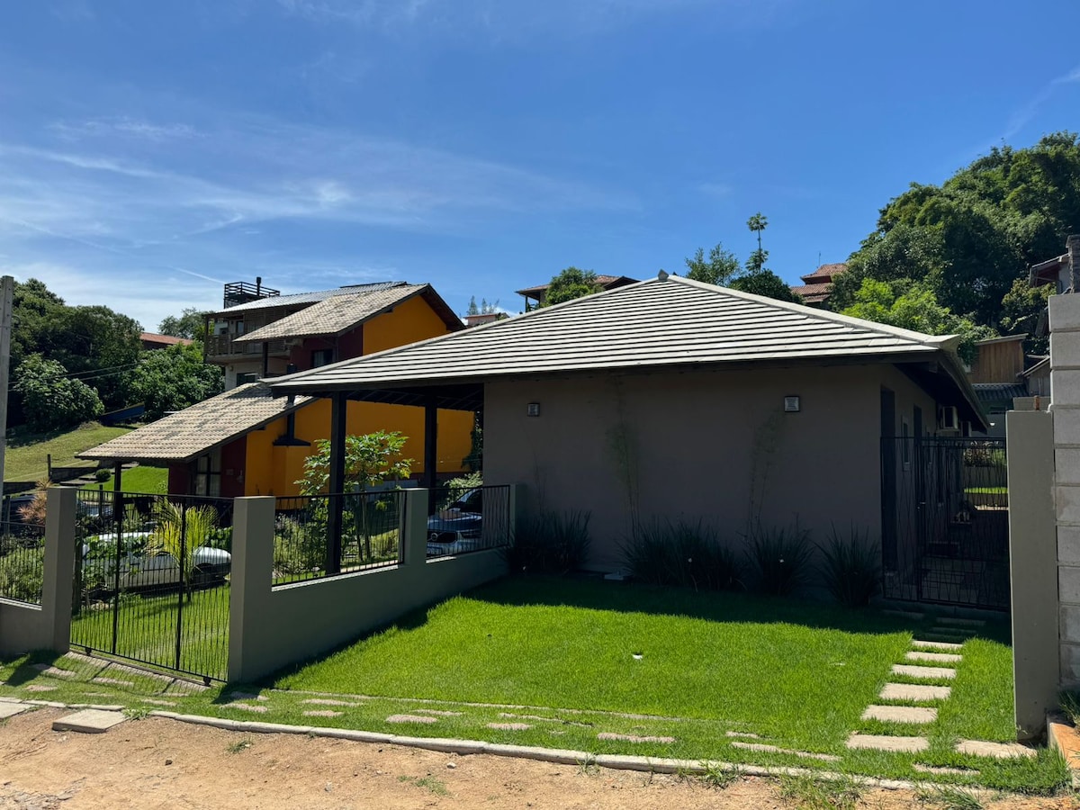 The exterior of a single-story house is presented, featuring a sloped roof and a manicured lawn. A pathway of stepping stones leads to the entrance. Surrounding vegetation and a gated fence enhance privacy, with other colorful homes visible in the background.