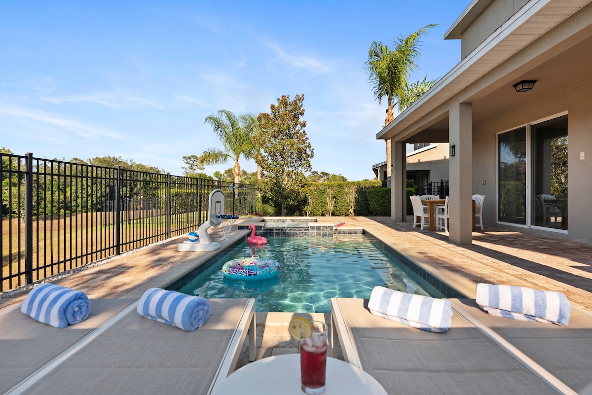 A private backyard features a heated pool and spa, surrounded by lounge chairs with striped towels. A refreshing drink sits on a table nearby, with palm trees visible in the background. The area is enclosed by a black safety fence, ensuring privacy.