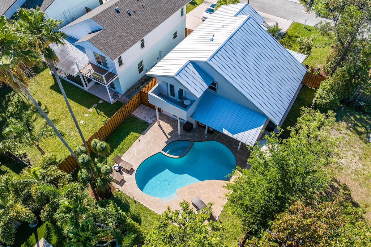 An aerial view captures a spacious backyard featuring a shaped swimming pool surrounded by patio space. Sun loungers are positioned along the edges, and neighboring properties are visible with green foliage enhancing the serene setting.
