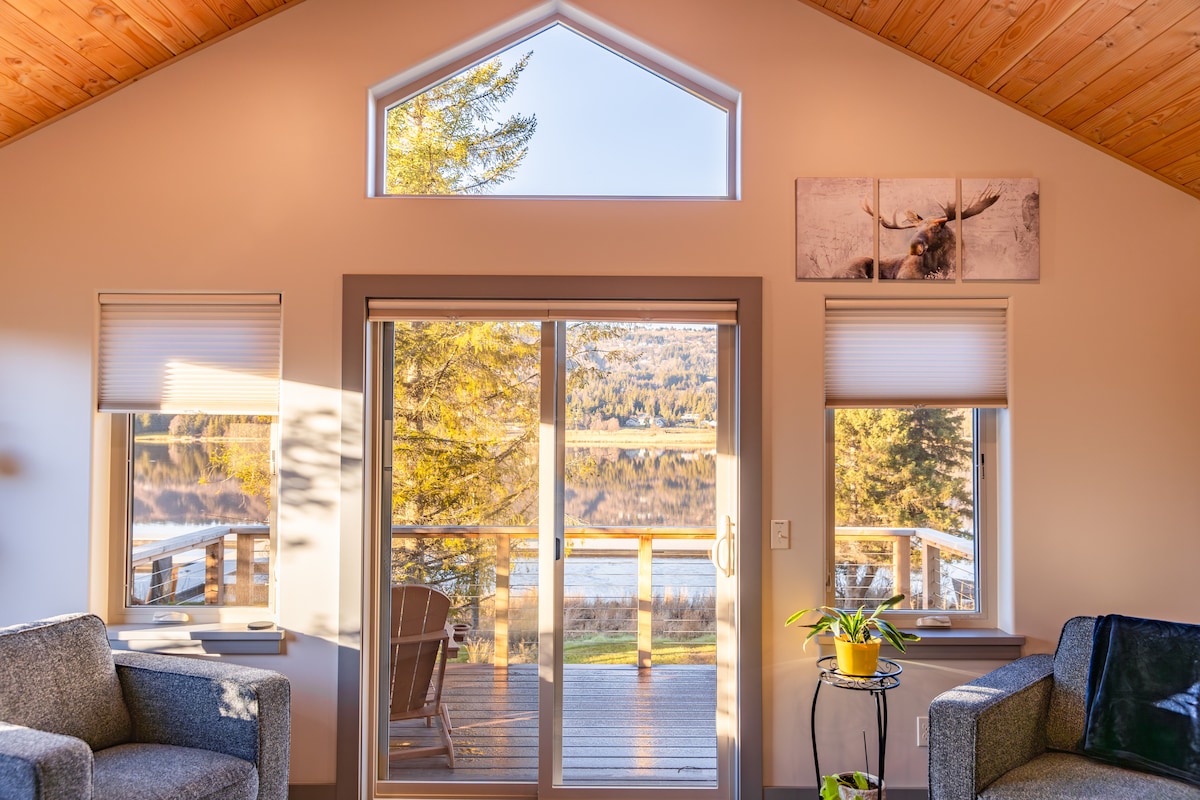 Large glass doors offer a view of the lakeside, framed by two comfortable seating areas. Natural light fills the space, highlighting the wooden ceiling and a serene atmosphere. A small potted plant adds a touch of greenery beside the window.
