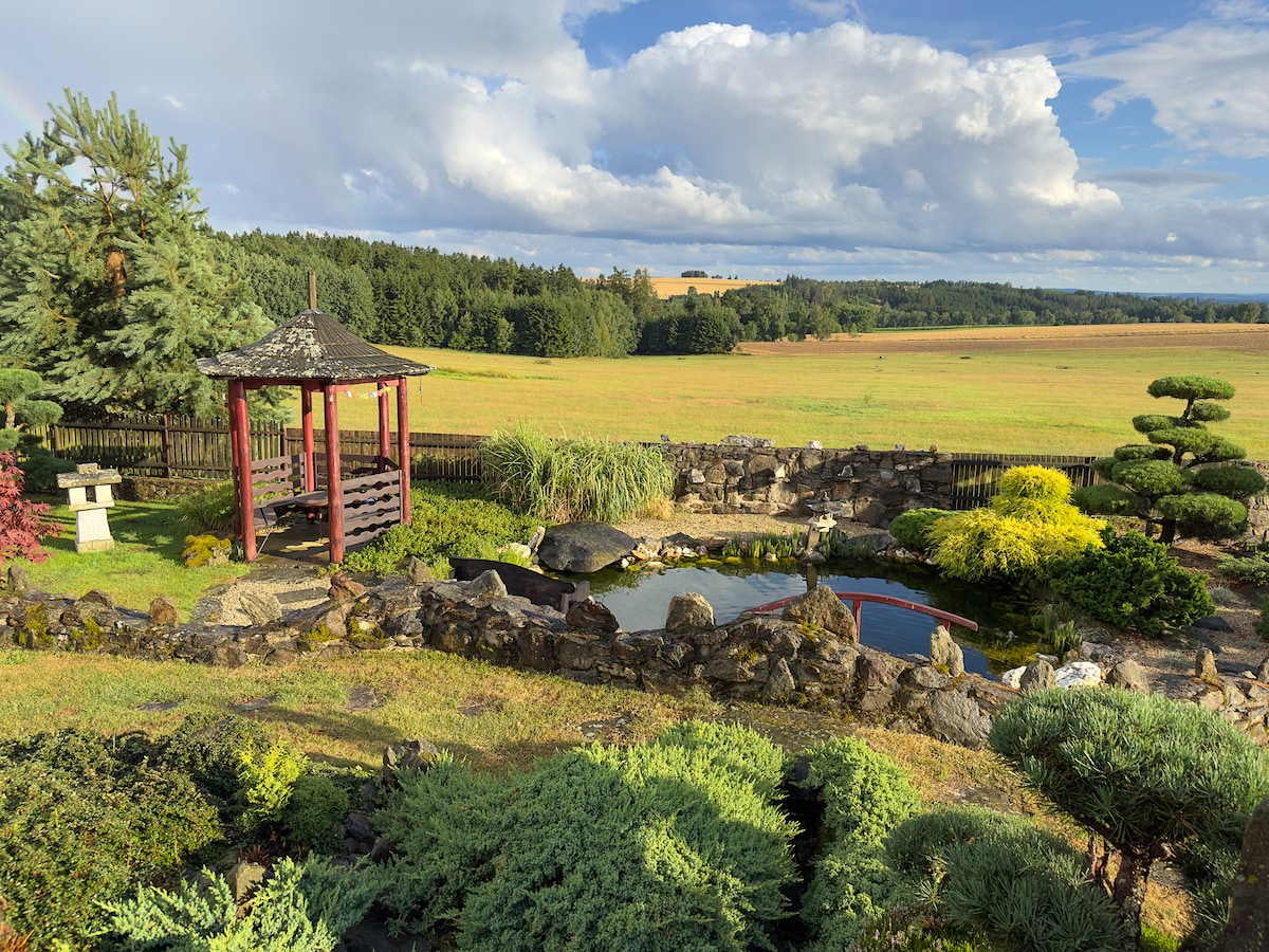 A serene Japanese-style garden features a koi pond surrounded by lush greenery and carefully shaped shrubs. A traditional gazebo stands nearby, offering a place to relax. The open landscape beyond showcases fields and dense forests under a partly cloudy sky.