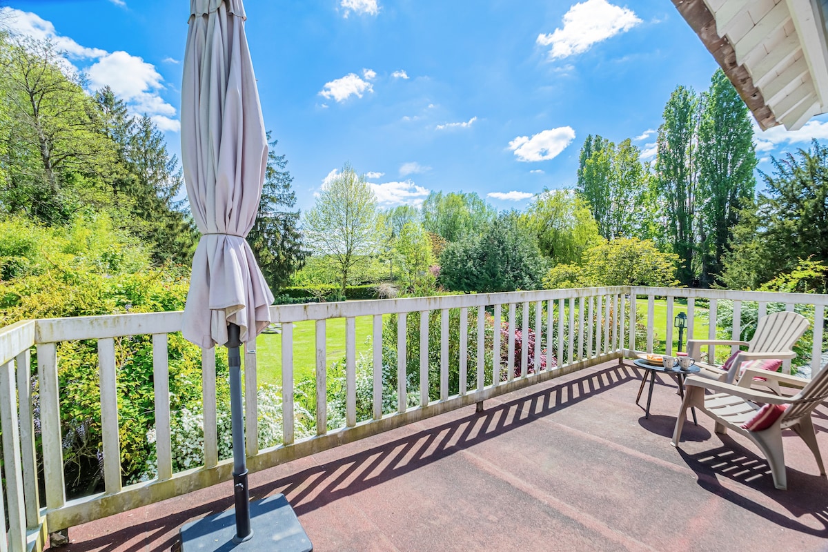 A spacious terrace is visible, featuring two lounge chairs and a small table. A large umbrella provides shade, while lush greenery and colorful flowers surround the area, contributing to a peaceful outdoor setting. The blue sky is dotted with fluffy white clouds, enhancing the serene atmosphere.