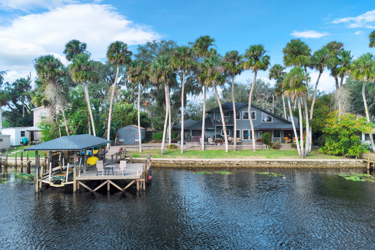 The home is situated along the St. John's River, featuring a spacious backyard with palm trees lining the property. A dock is visible, equipped with seating, providing a serene spot for relaxation. The architecture displays a two-story structure, enhancing the appeal of the riverside location.