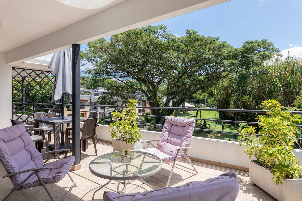 A spacious terrace features comfortable seating arrangements, including two lounge chairs and a dining table. Lush greenery is visible in the background, providing a serene view. Natural light enhances the space, creating an inviting environment for relaxation or gatherings.