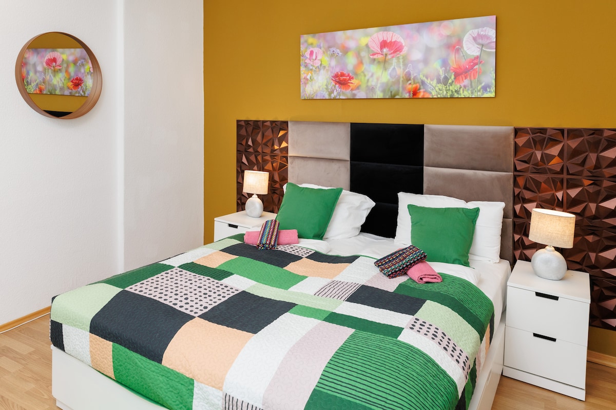 A spacious bedroom features a large bed with a multicolored patchwork quilt and green accent pillows. Two bedside lamps provide soft lighting. A round mirror and a floral artwork in vibrant colors adorn the walls, enhancing the room's contemporary design.